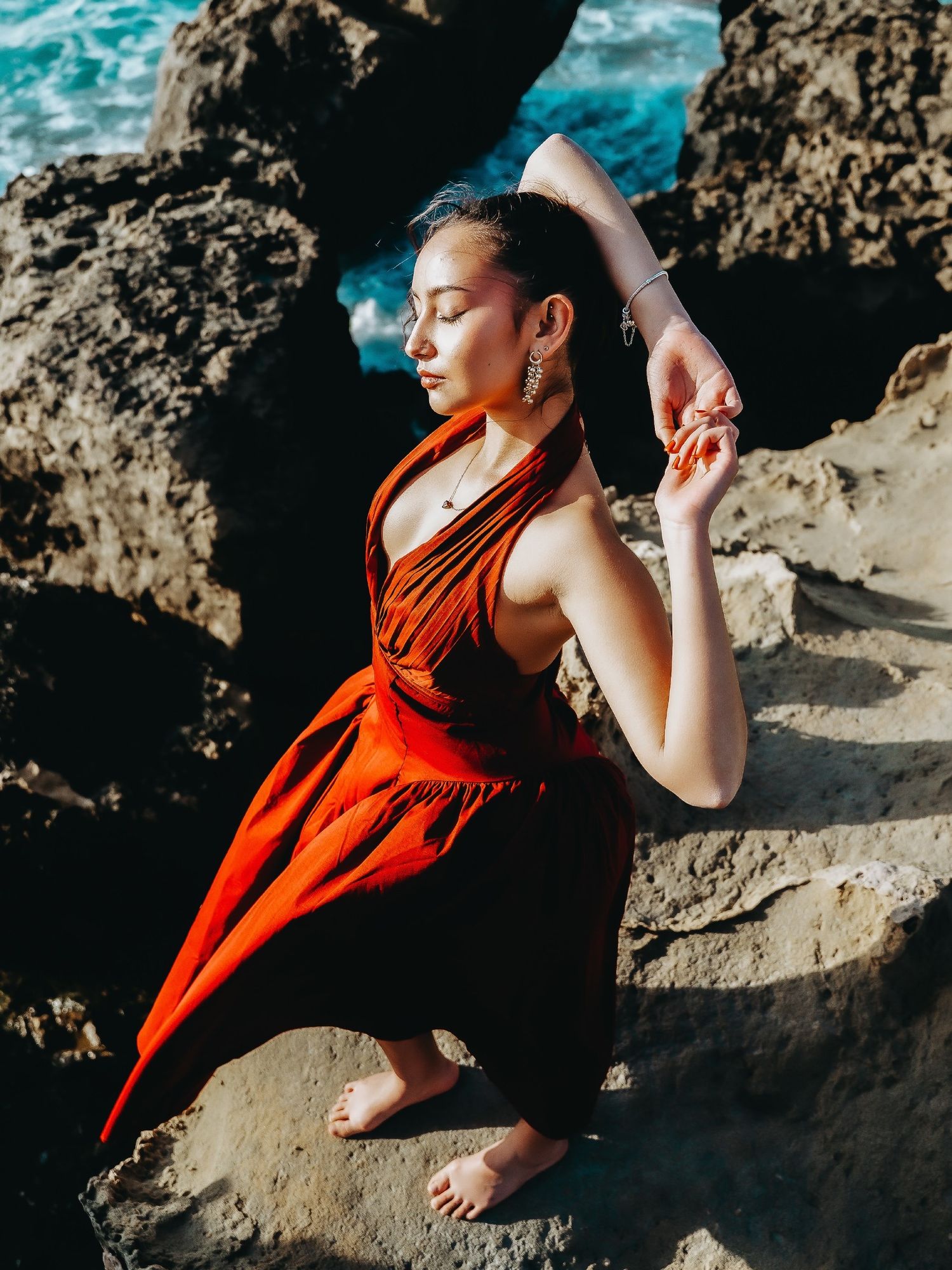 Woman in red dress practicing yoga on rocky coastal cliffs with ocean waves in the background.