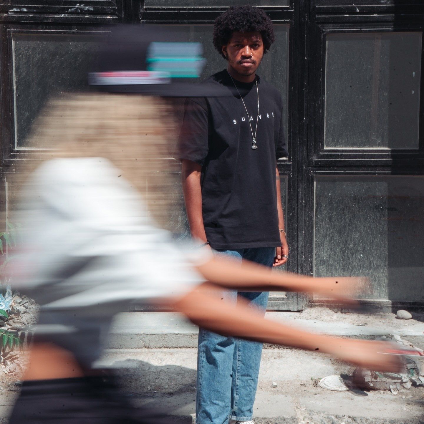 A young man in a black oversized tee and jeans stands still as a blurred figure moves past him on a city sidewalk.