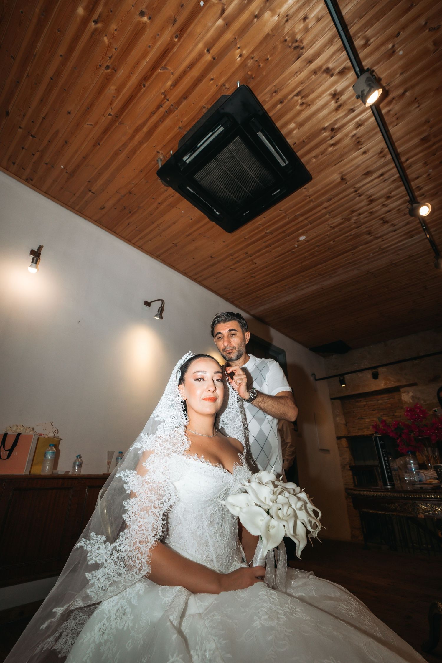 Bride in white lace gown and veil holds bouquet while groom stands behind her in a warmly lit wooden venue.