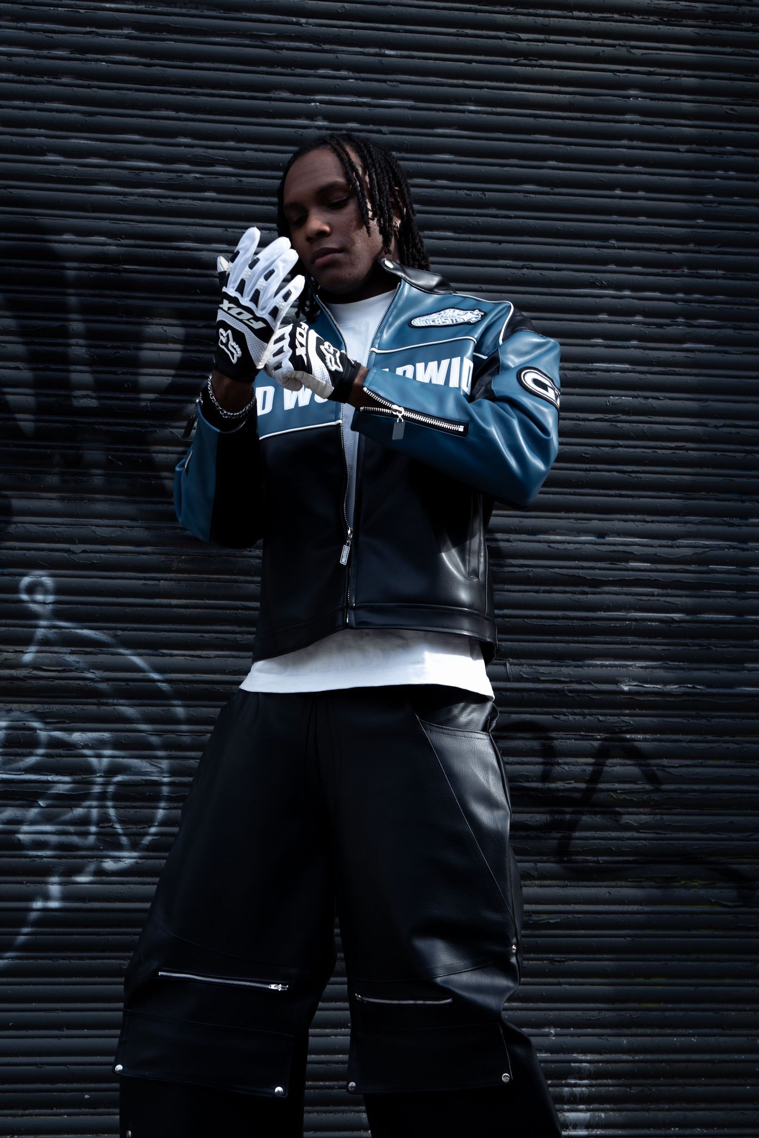 Young man in teal leather moto jacket, white gloves, and black leather pants posing against dark metal wall with graffiti.
