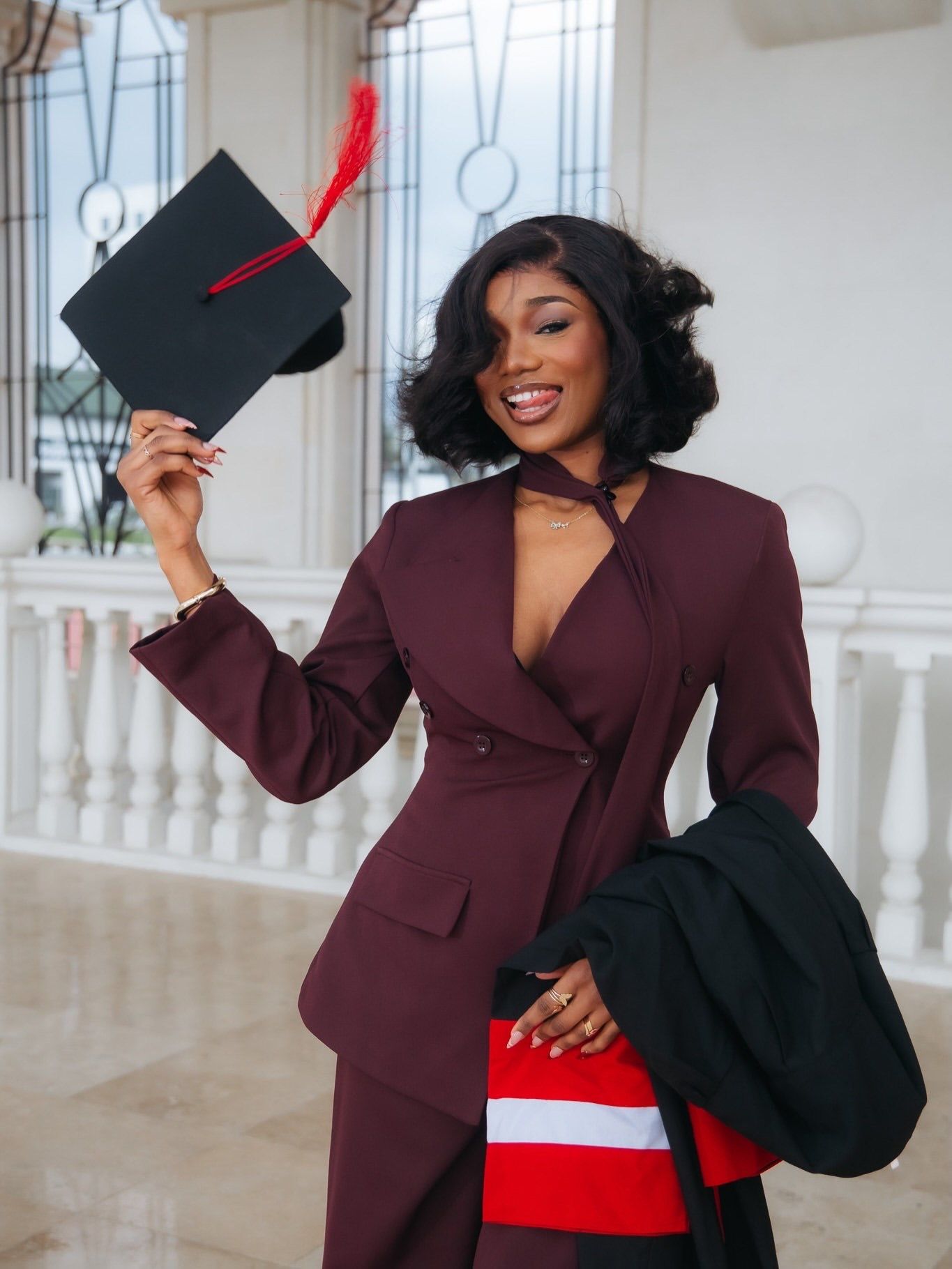 Joyful Black woman in burgundy suit holds graduation cap with red feather tassel indoors.