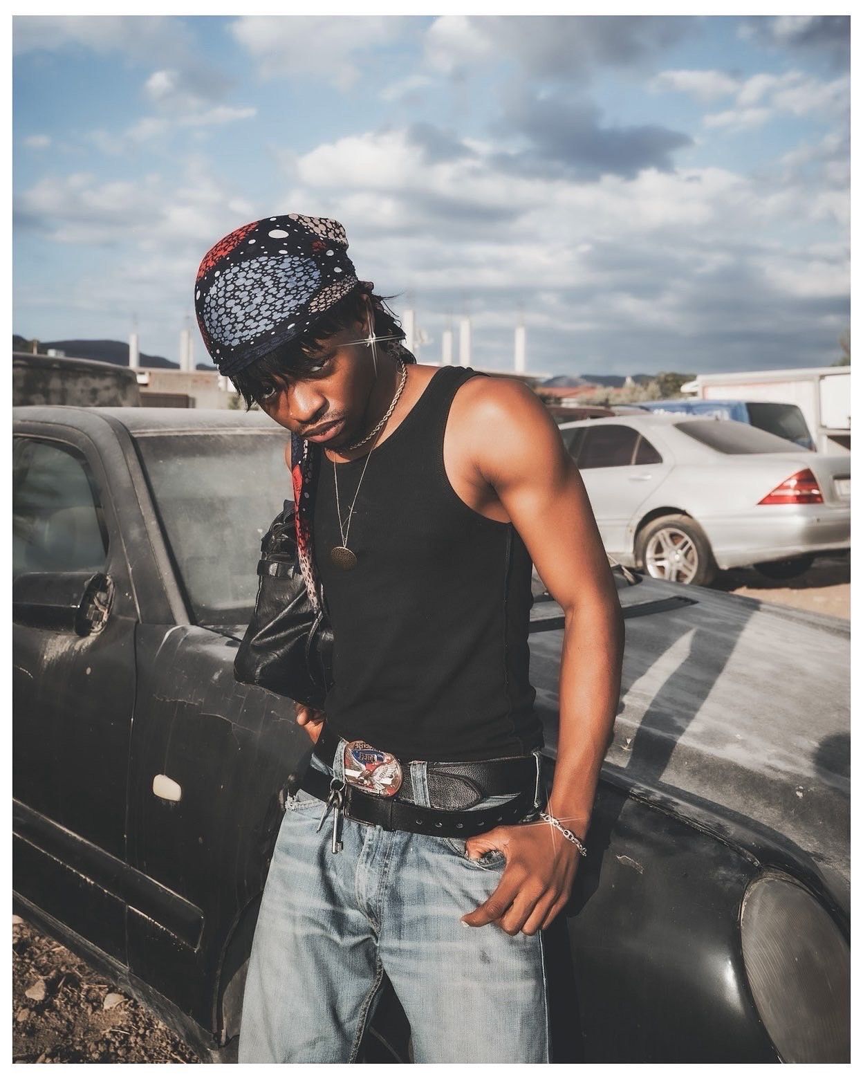 Young man in black tank top, jeans, and red bandana poses leaning on a car in a parking lot.