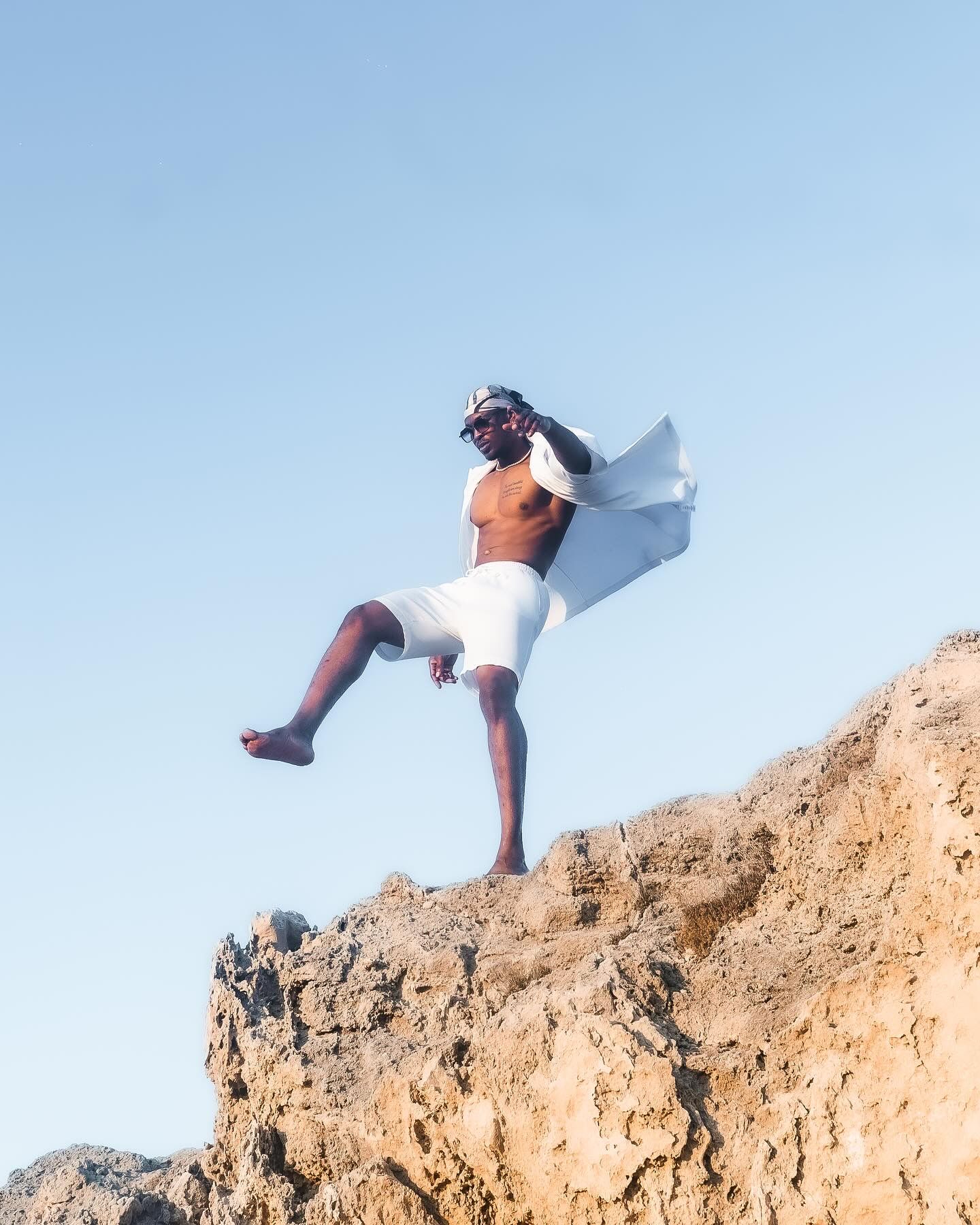 A shirtless man in white shorts stands triumphantly on a rocky cliff, cape flowing in the wind against a clear blue sky.