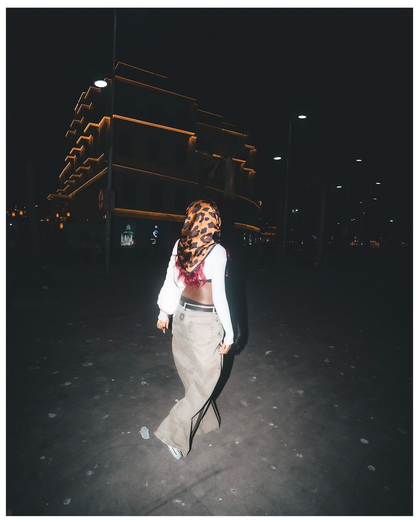 A woman wearing a hijab, white top, and beige wide-leg pants walks outdoors at night near an illuminated building.