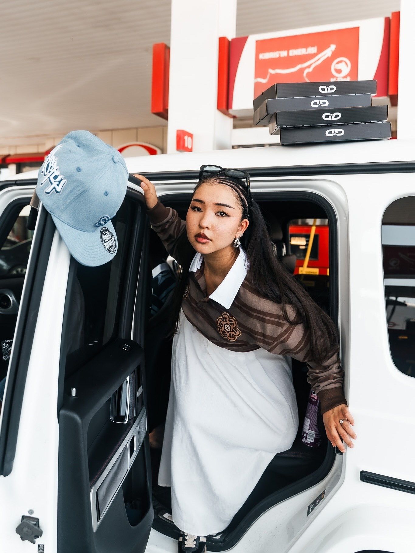Young woman in white dress and brown mesh top holds a blue cap while exiting a white SUV at a gas station.