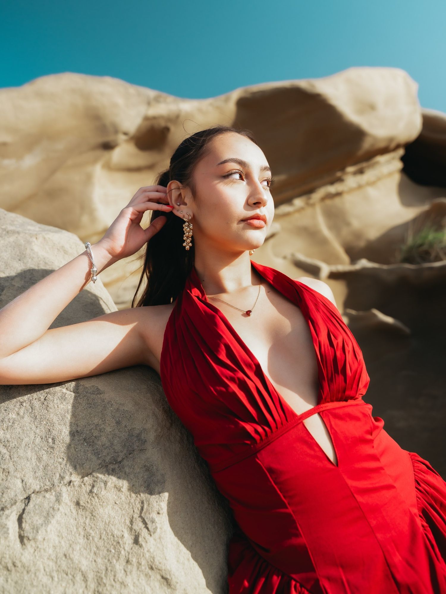 Woman in red halter dress leaning on sandstone rocks under blue sky.