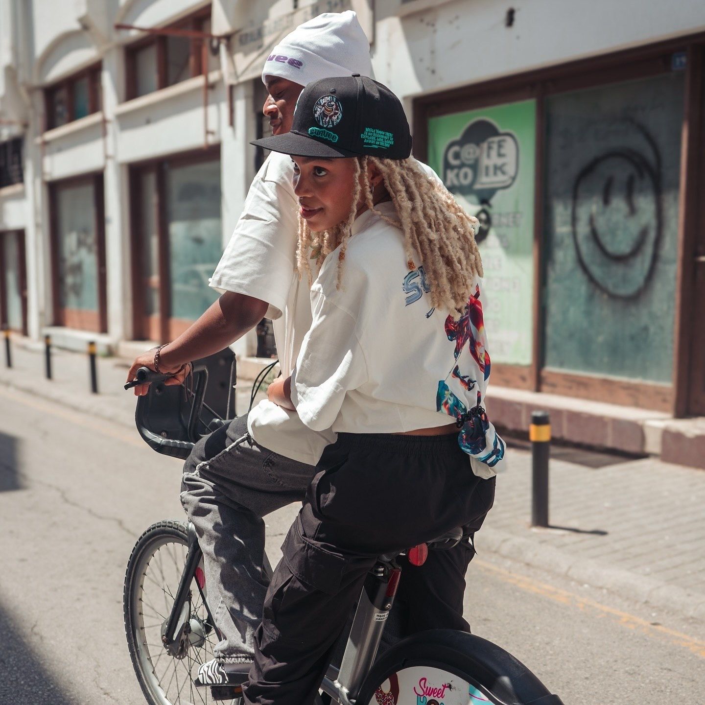 Two friends riding a bicycle together on a urban street, wearing casual streetwear and snapback hats.