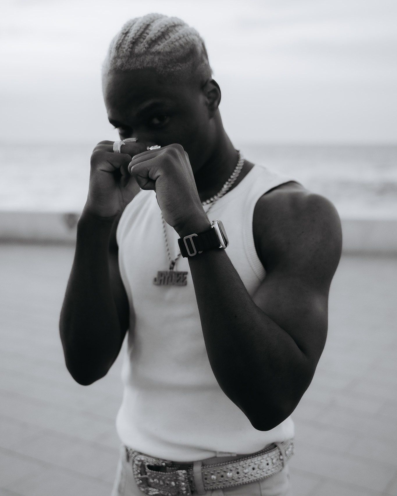 Muscular man in white tank top posing in boxing stance on beach, wearing chain necklace and watch, black and white photo.