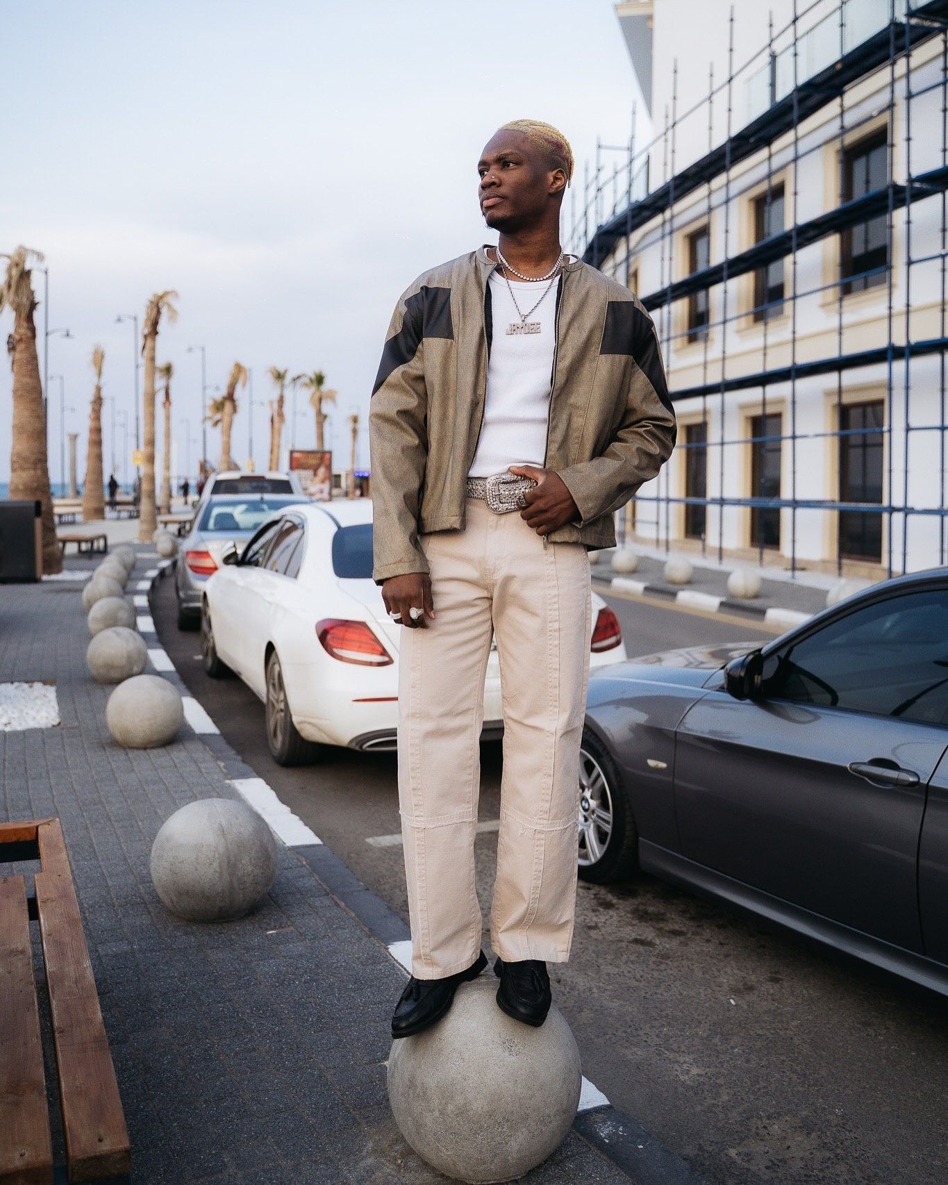 Stylish man standing on concrete sphere outdoors, wearing cream pants, olive jacket, and white tee near parked cars.