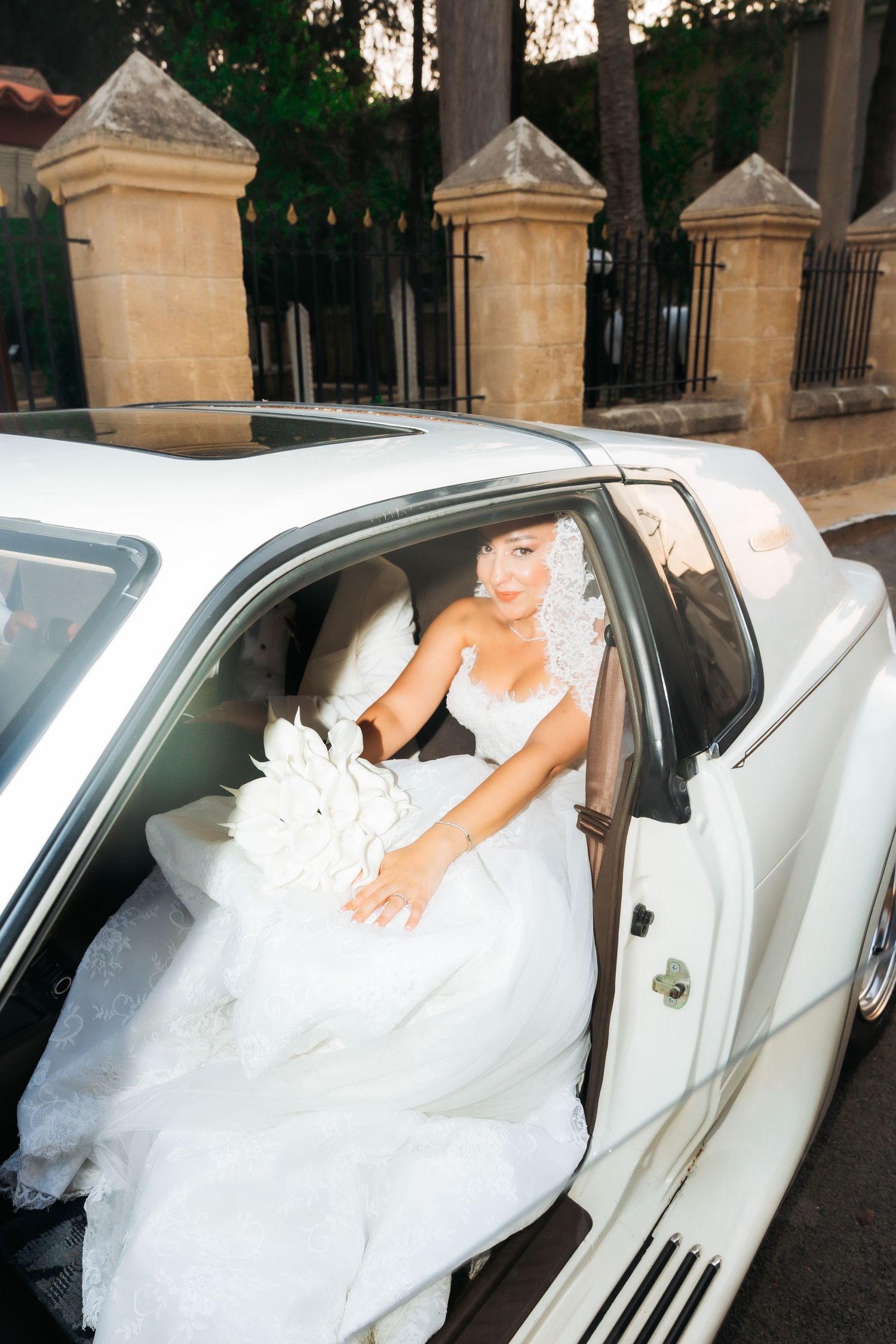 Bride in white gown holding floral bouquet stepping out of a white luxury limousine on her wedding day.