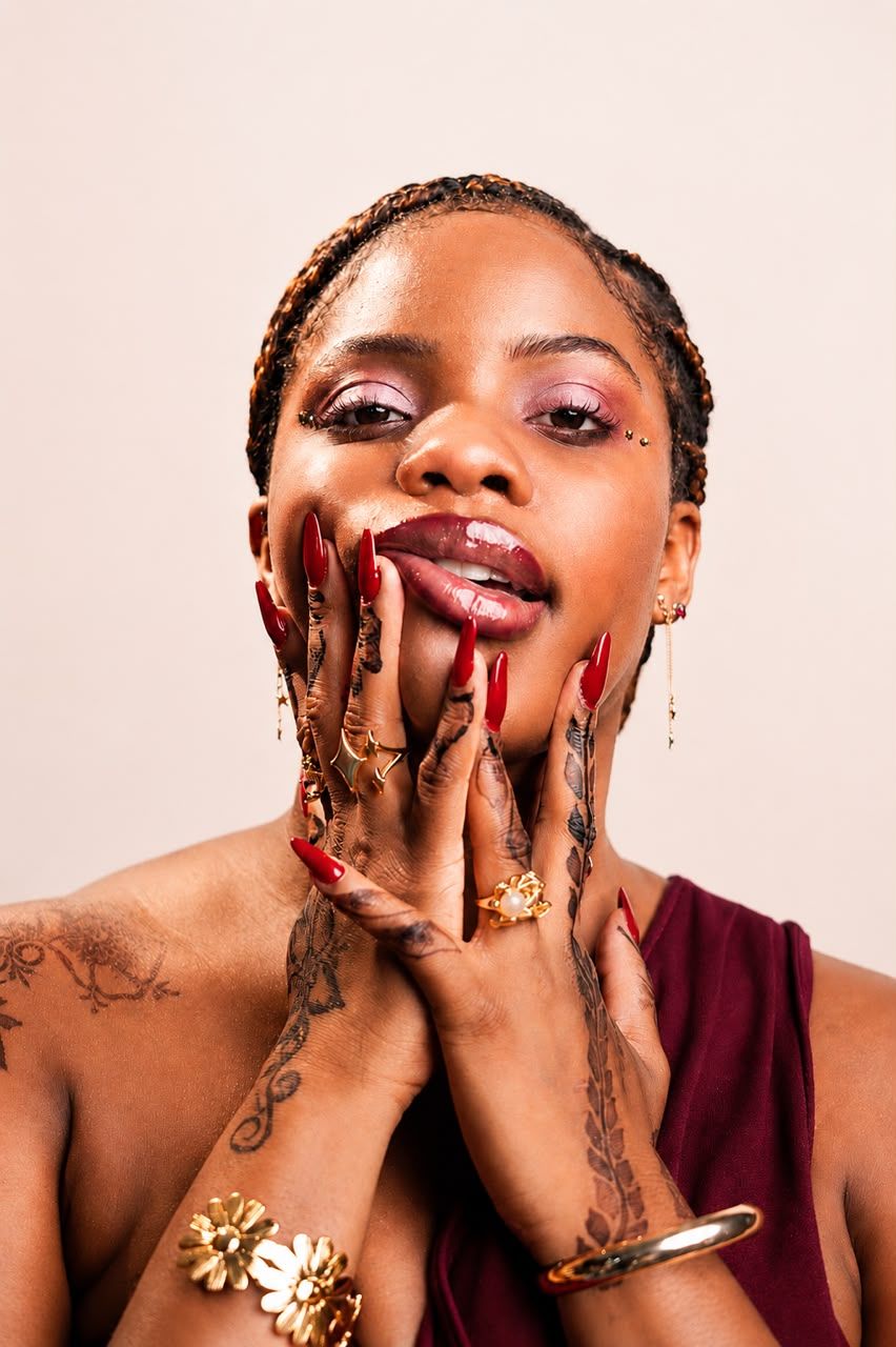 A Black woman with braids, red nails, tattoos, and gold jewelry poses gracefully with hands framing her face.