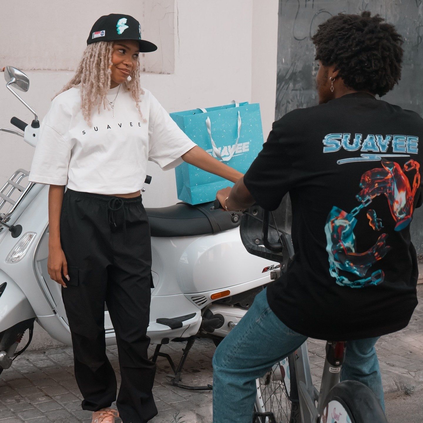 Two friends pose outdoors near a scooter, wearing stylish streetwear including graphic tees and cargo pants.