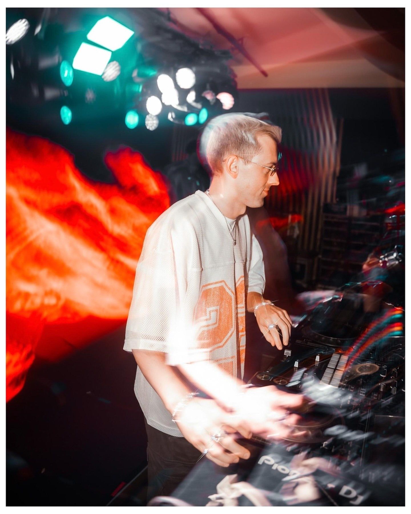 A DJ with blonde hair performs at a nightclub, working the decks under colorful stage lighting.