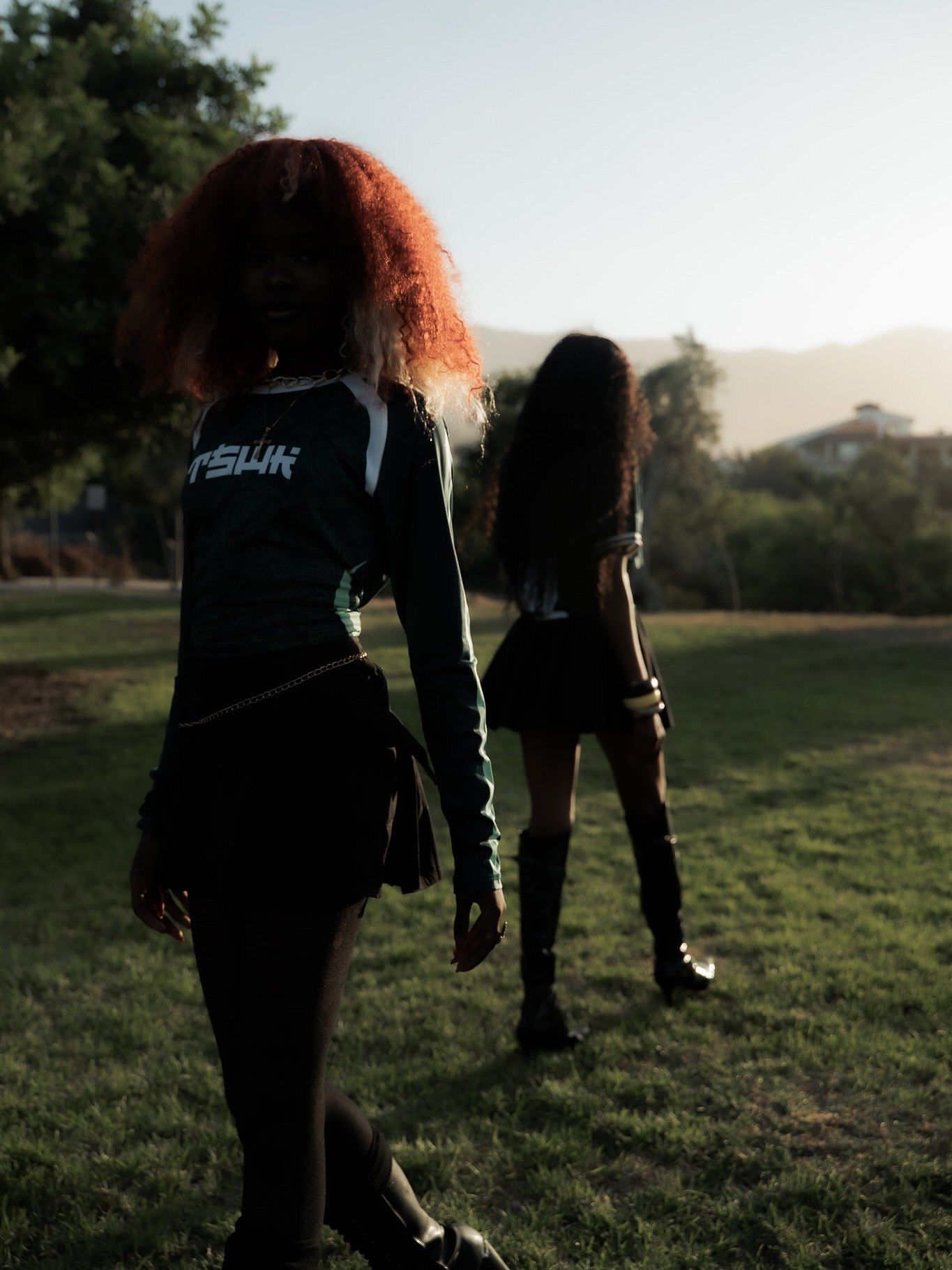 Two women in dark streetwear outfits stand on a grassy field at golden hour, one with bold red curly hair.