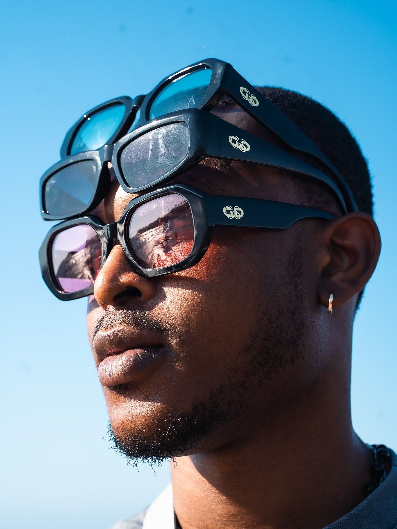 Close-up portrait of a man stacking multiple black designer sunglasses on his face against a blue sky.