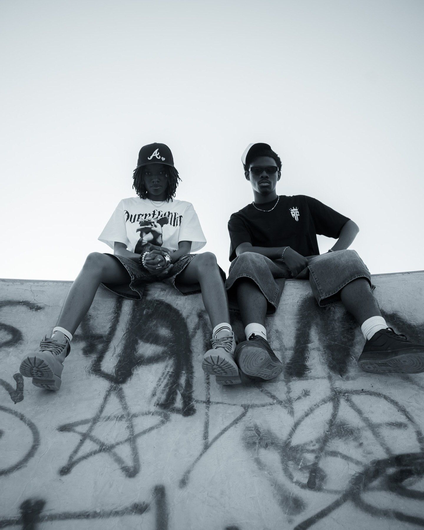 Two teens sit on a graffiti-covered skate ramp wearing streetwear in a black and white photo.