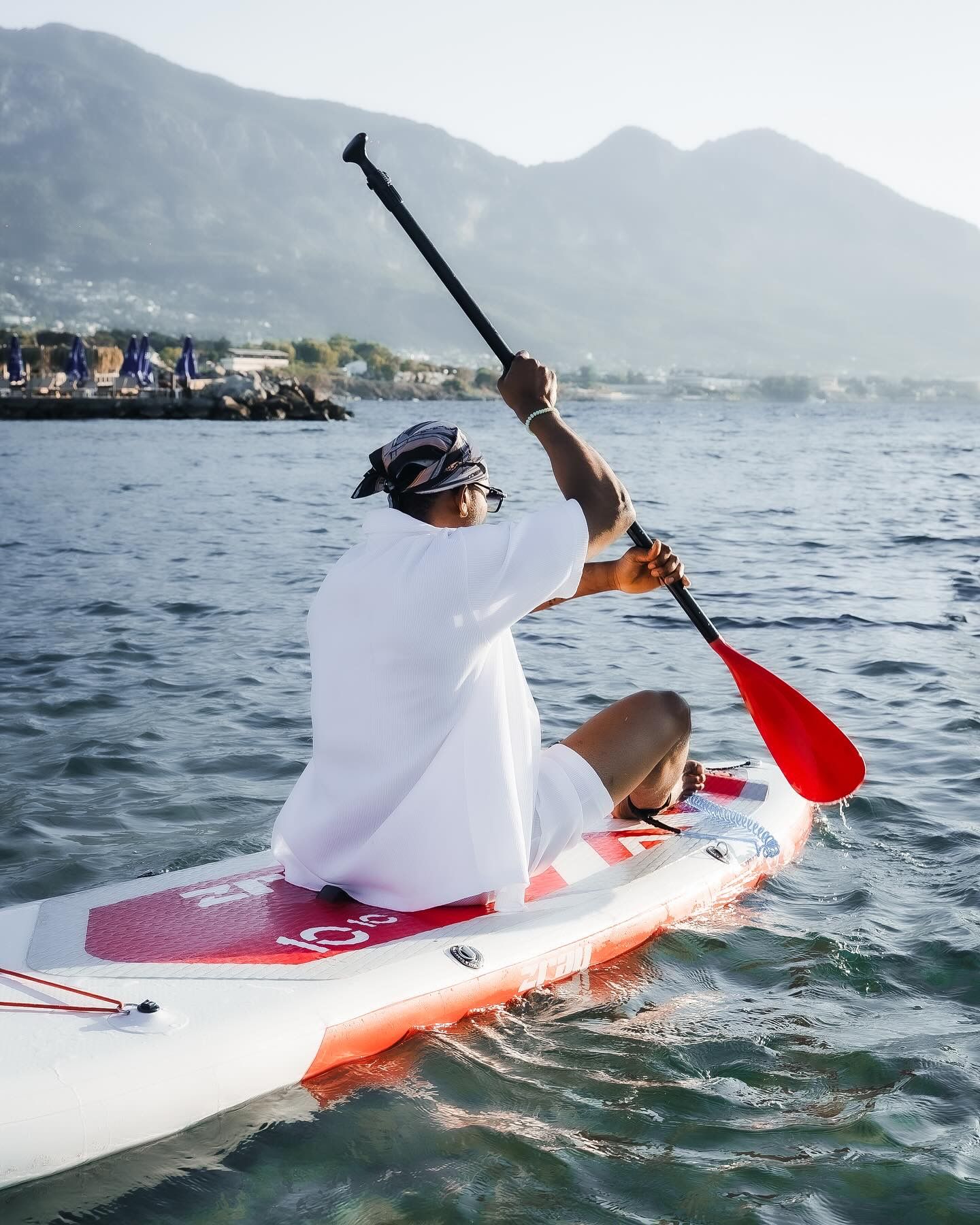 Person in white outfit paddling on a red SUP board on calm water with mountains in the background.