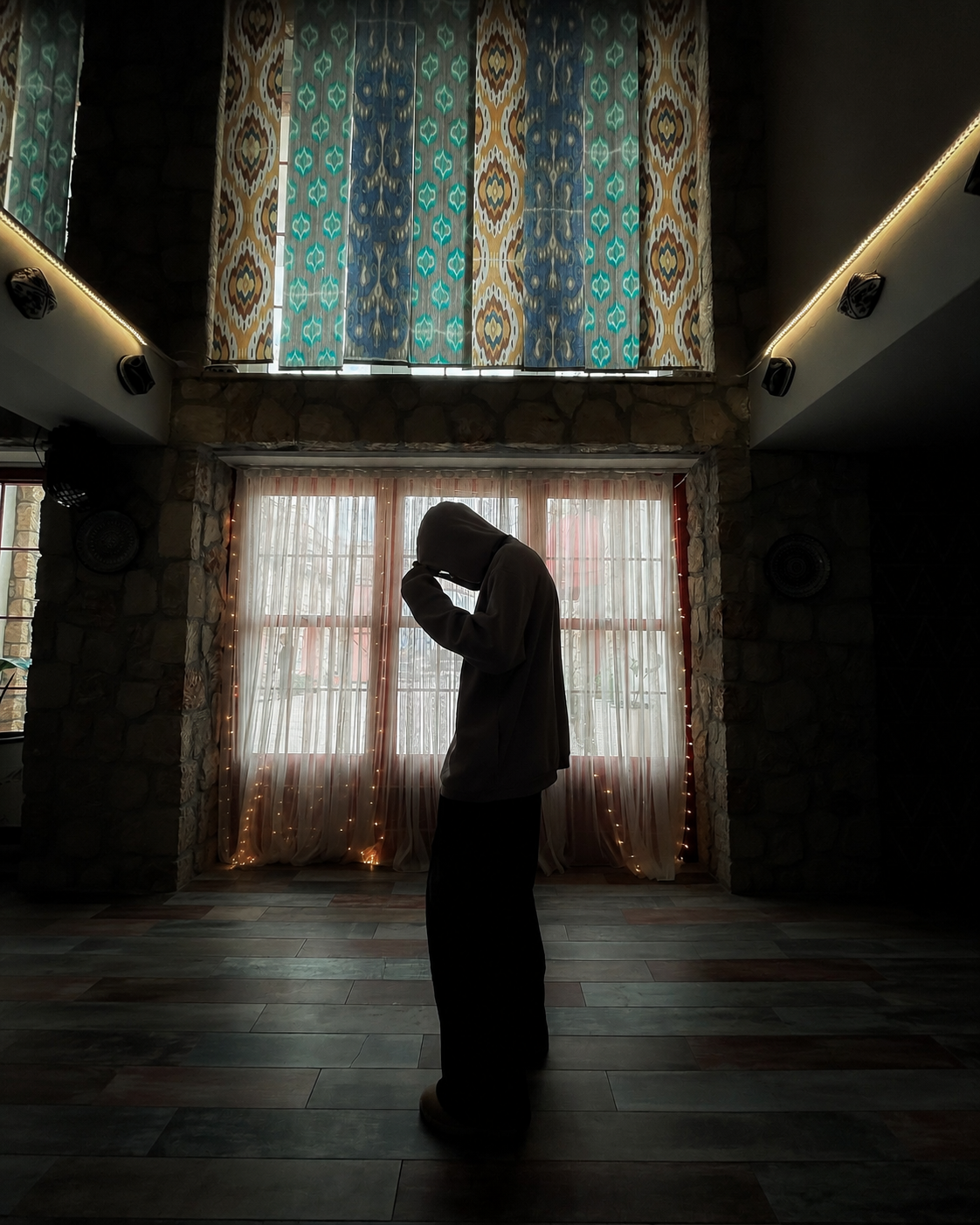 Silhouette of a person praying in a dimly lit room with ornate stained glass windows and sheer curtains.