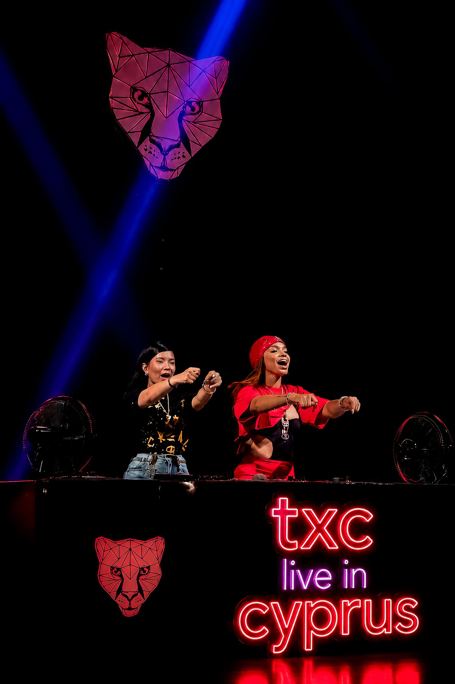 TXC duo performing live on stage in Cyprus, with neon sign reading 'txc live in cyprus' and geometric cat logo.