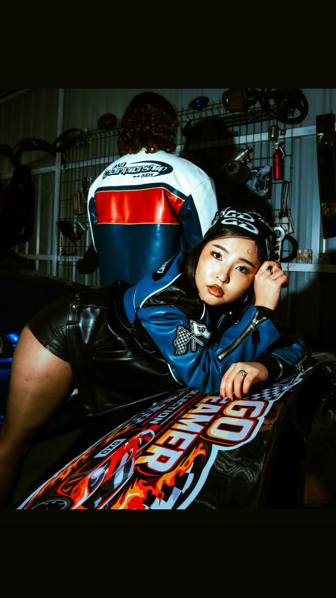 Young woman in blue racing jacket poses with motorcycle helmet in a garage setting.