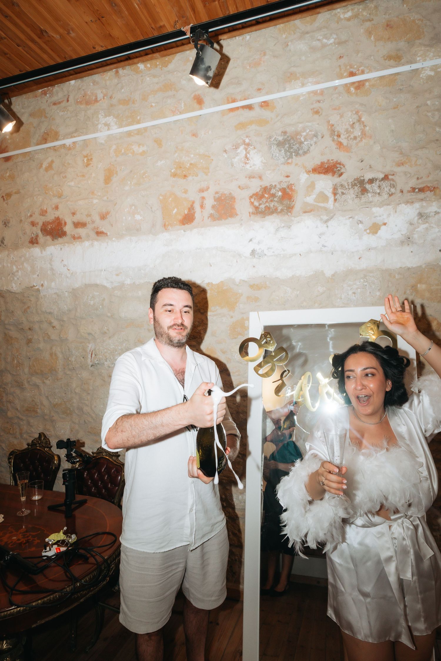 Couple celebrating with champagne and sparklers in a rustic stone-walled venue, dressed in white party attire.
