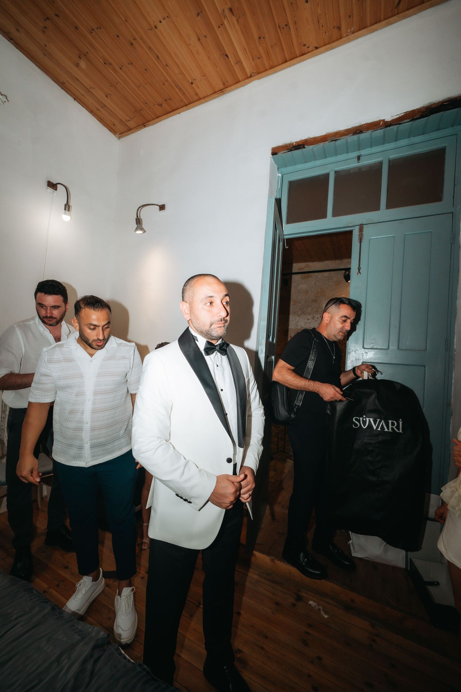 Groom in white tuxedo jacket with bow tie prepares with groomsmen in a room before wedding ceremony.