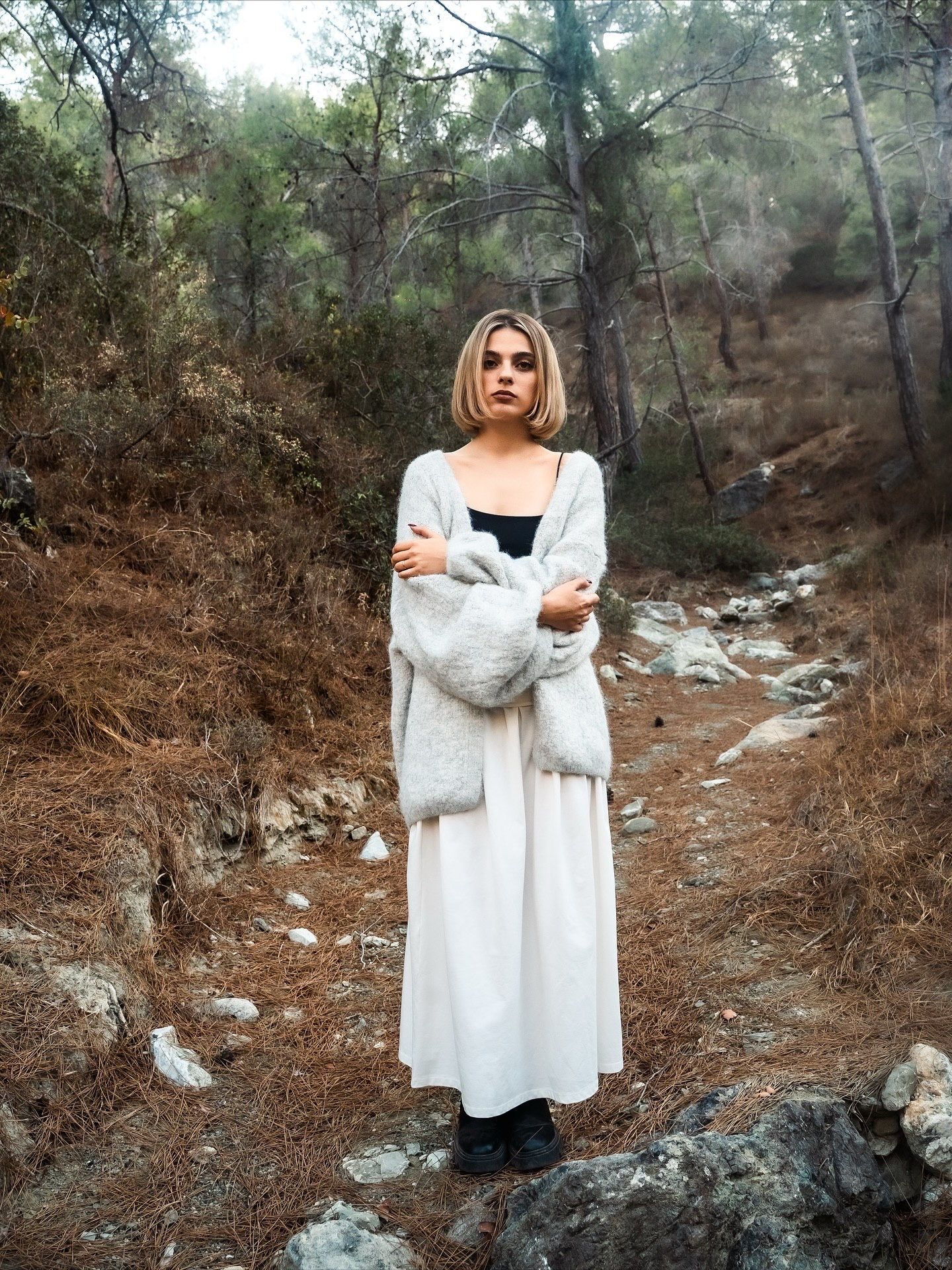 Young woman in white maxi dress and gray fur coat standing on rocky forest path among pine trees.