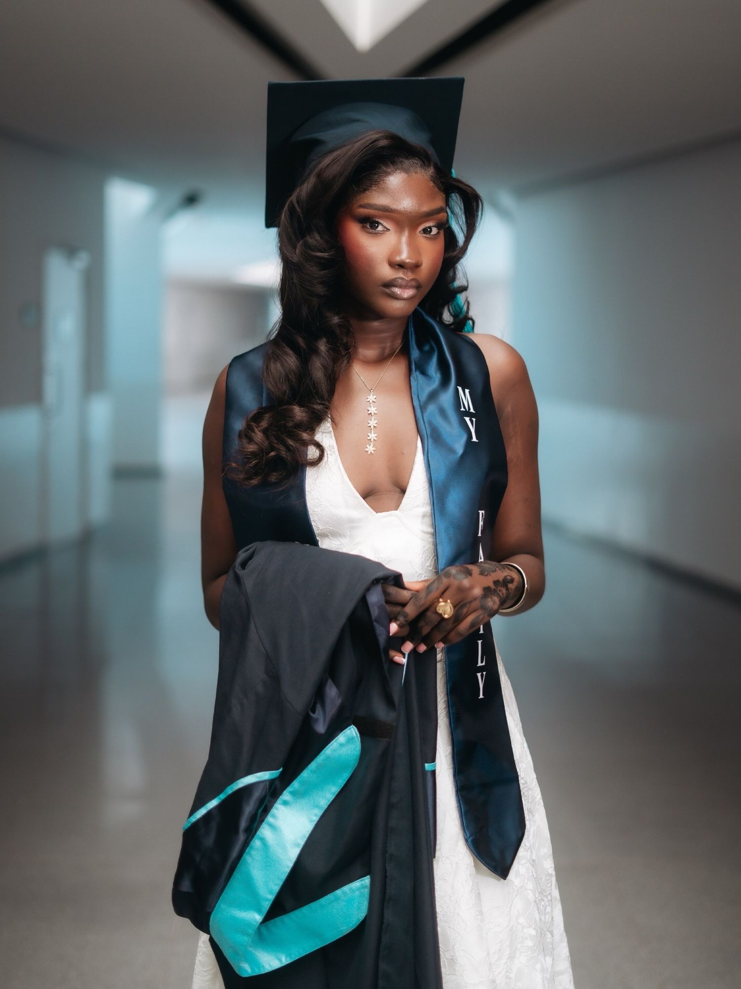 Graduate in white dress holding cap and gown with teal trim in a hallway.