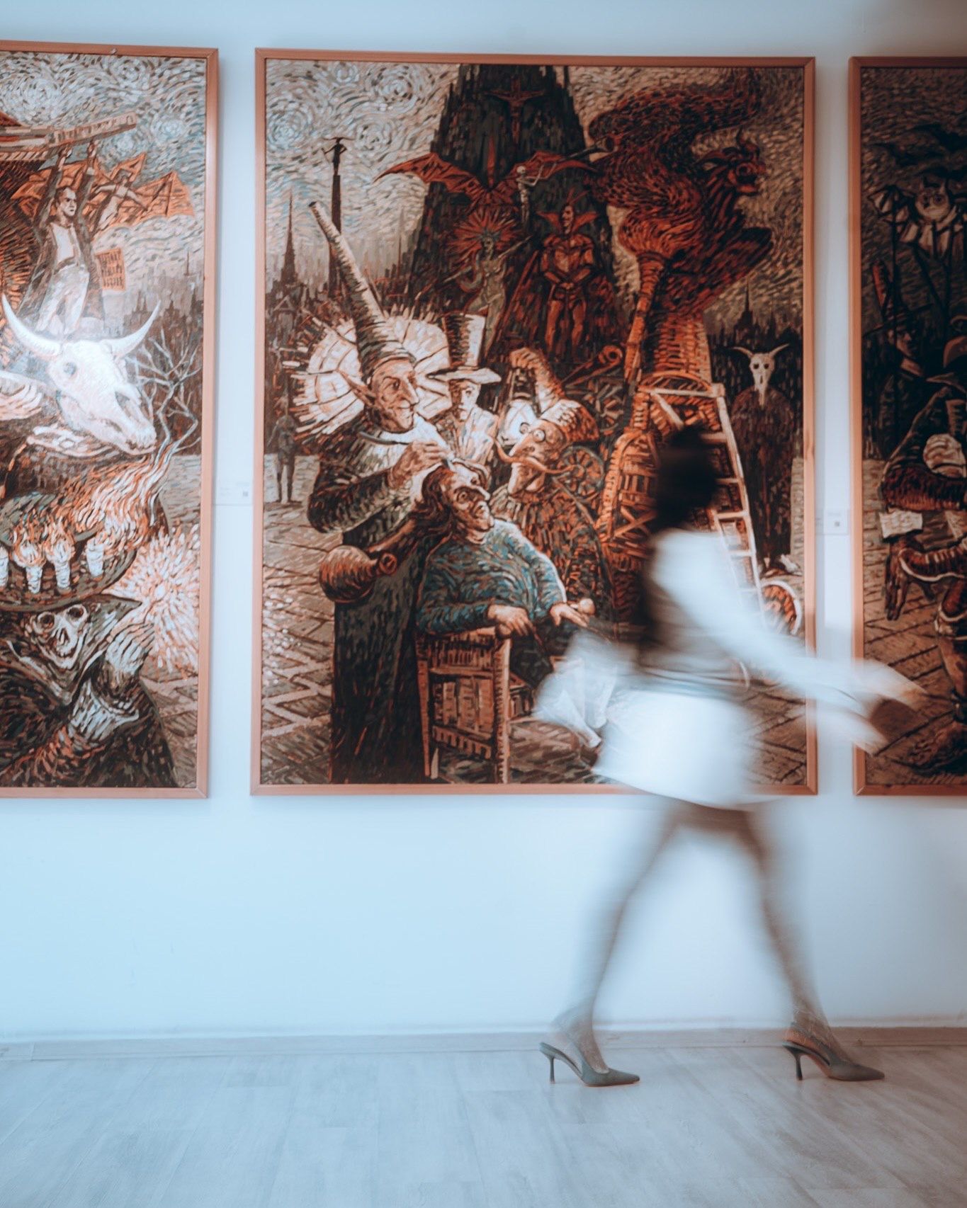 A blurred woman in white walks past large ornate tapestry paintings in a modern gallery.