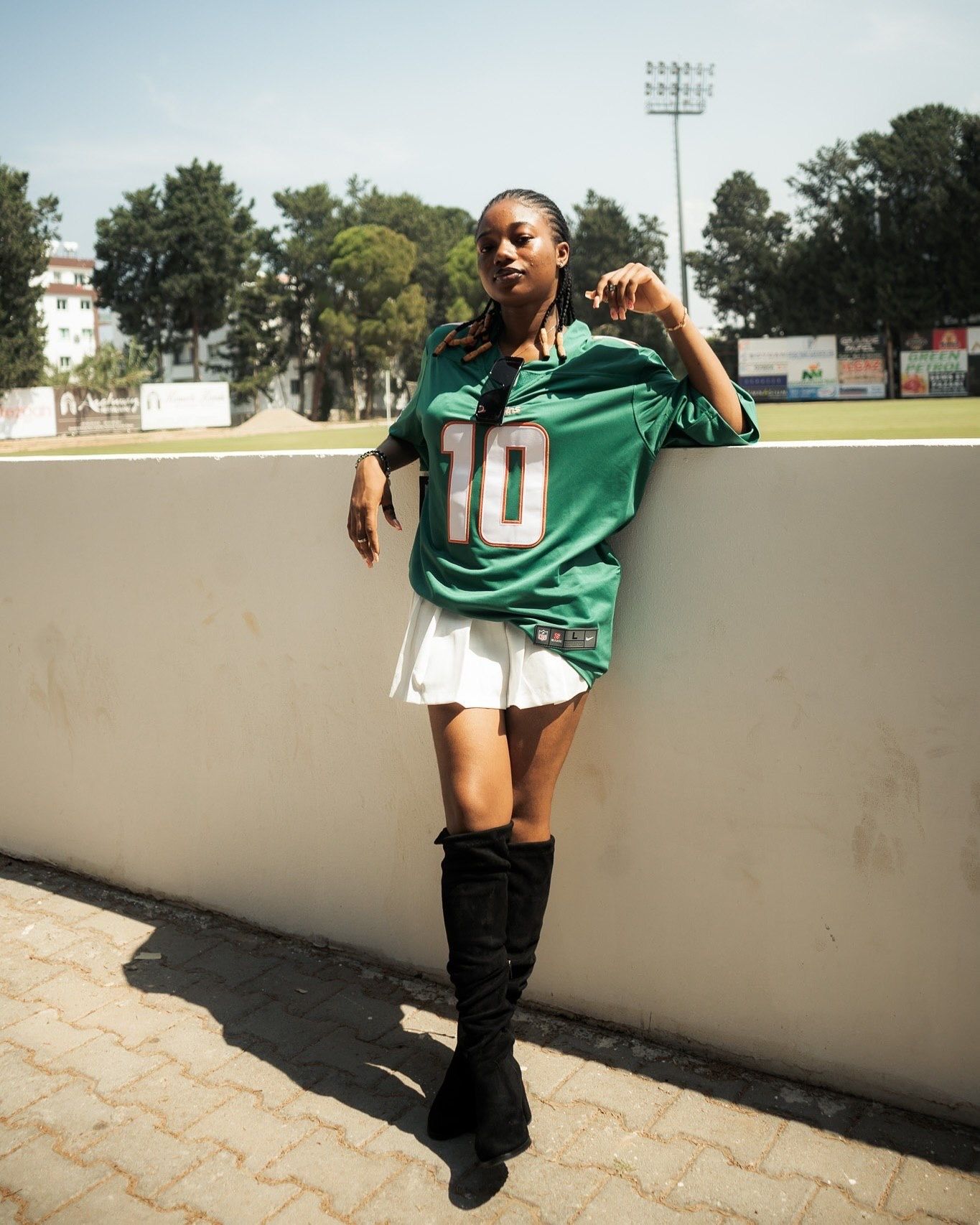 Woman in green #10 football jersey, white skirt, and black knee-high boots posing at a stadium.