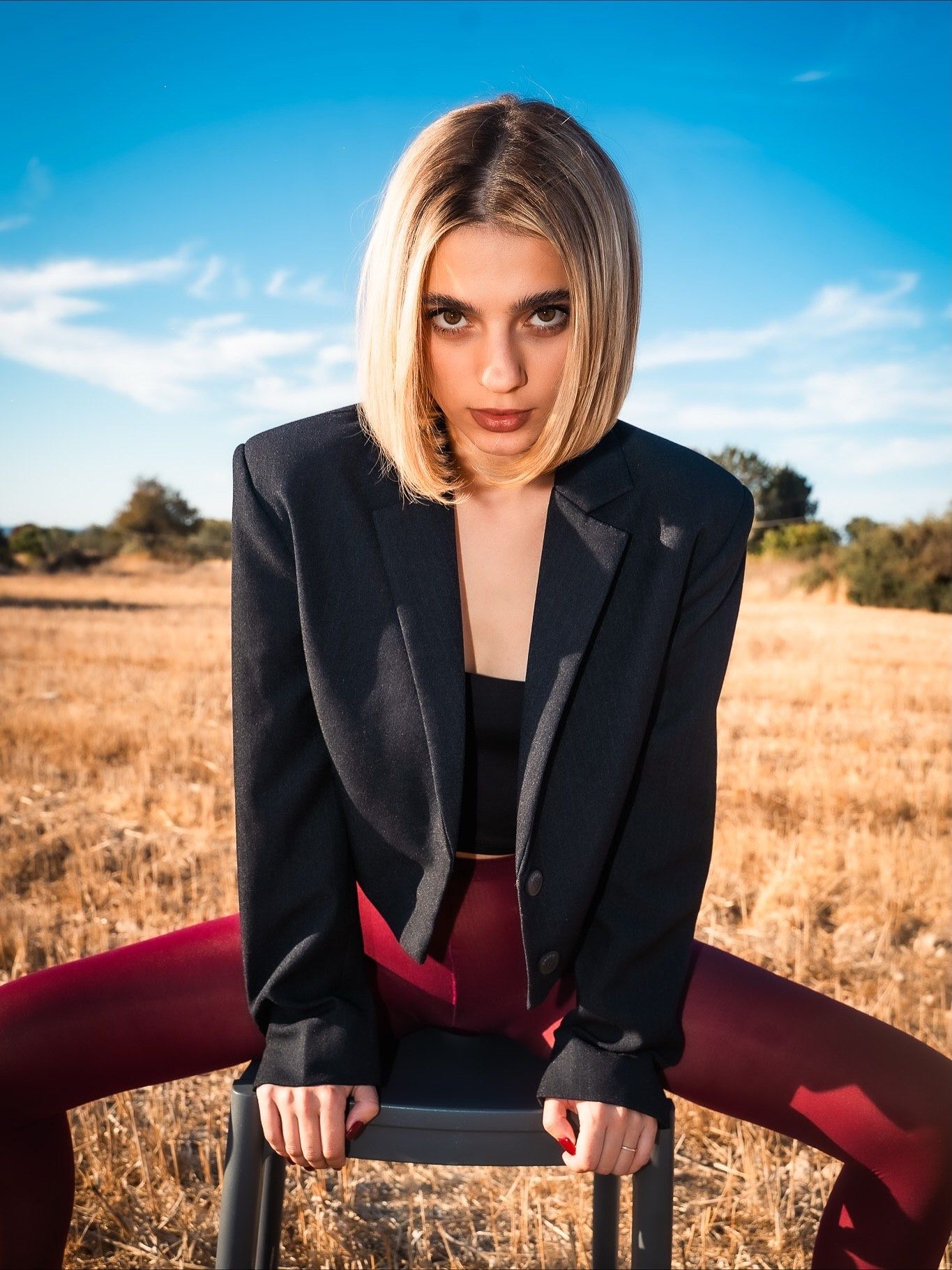 A young woman with a blonde bob poses in a black blazer and red pants, leaning on a chair in a golden field.