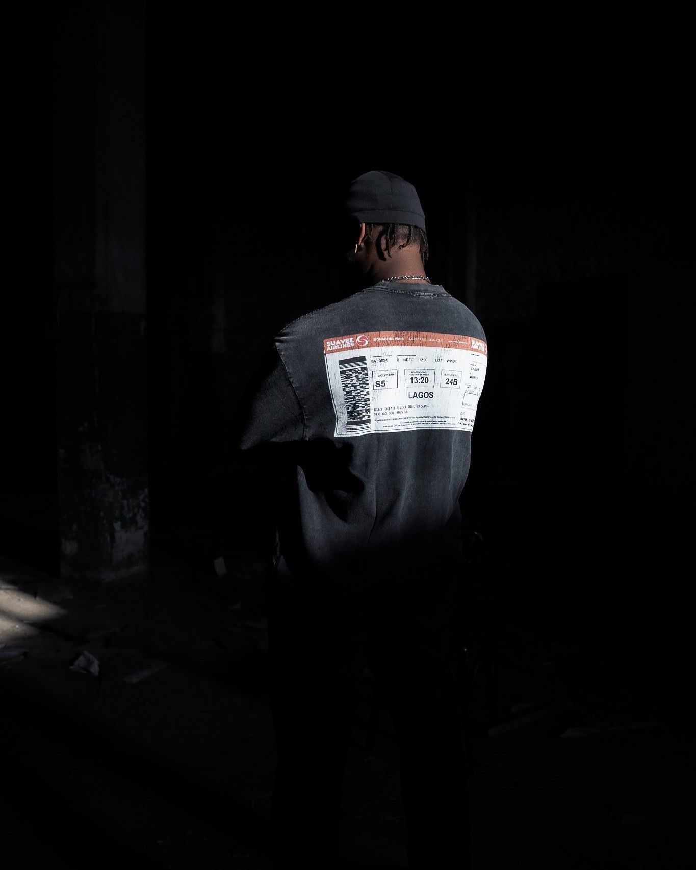 Person wearing dark oversized sweatshirt with printed label design, standing with back to camera in shadowy setting.