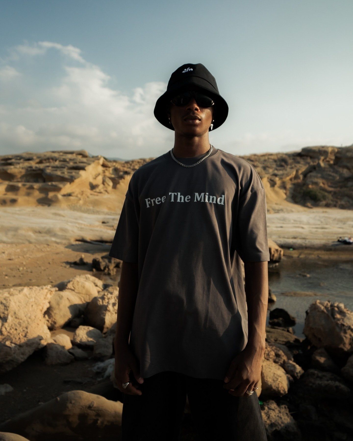 Man in black bucket hat and gray 'Free The Mind' t-shirt standing in rocky desert landscape.