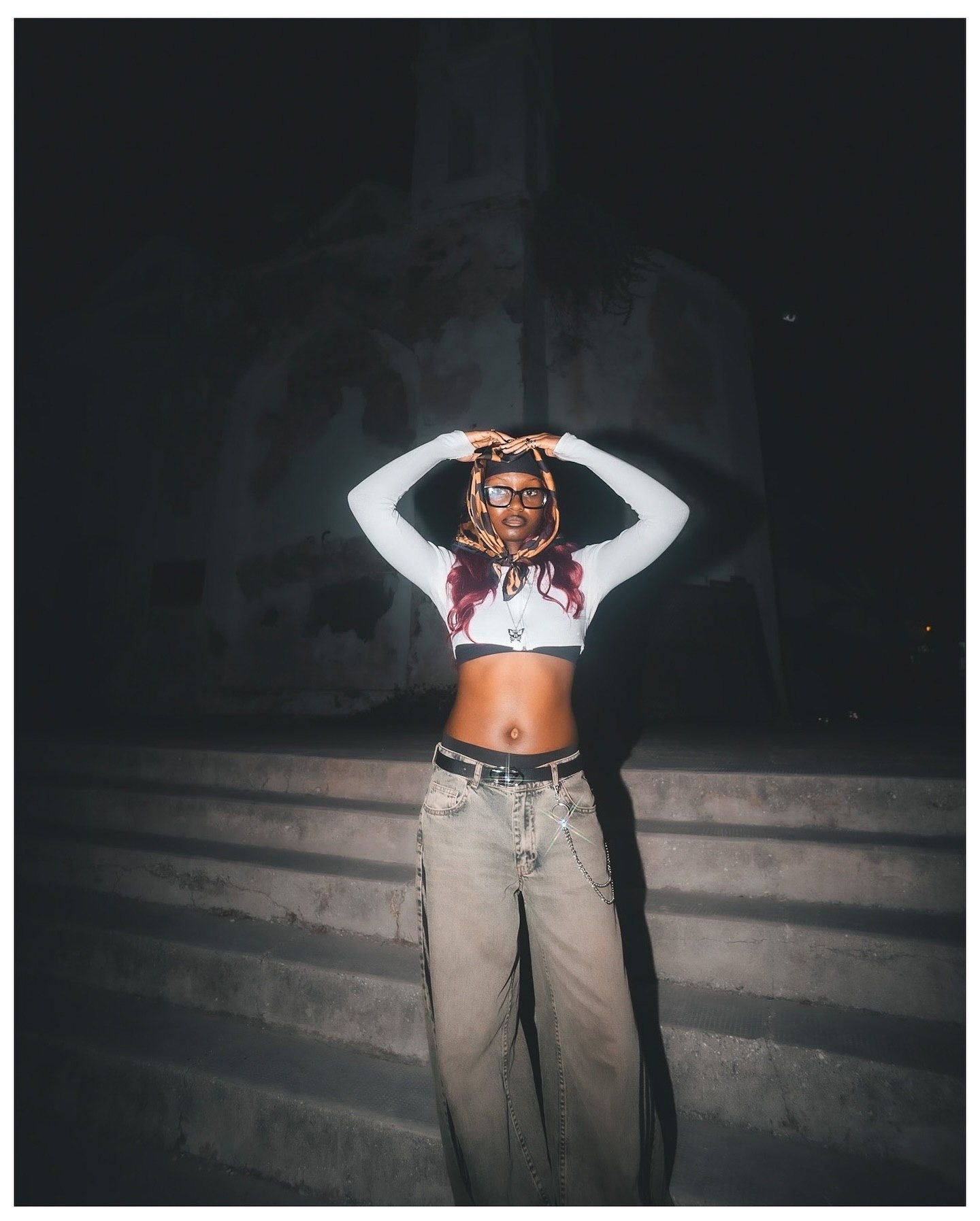 A young woman in a white crop top and baggy cargo jeans poses with hands on head under dramatic night lighting.