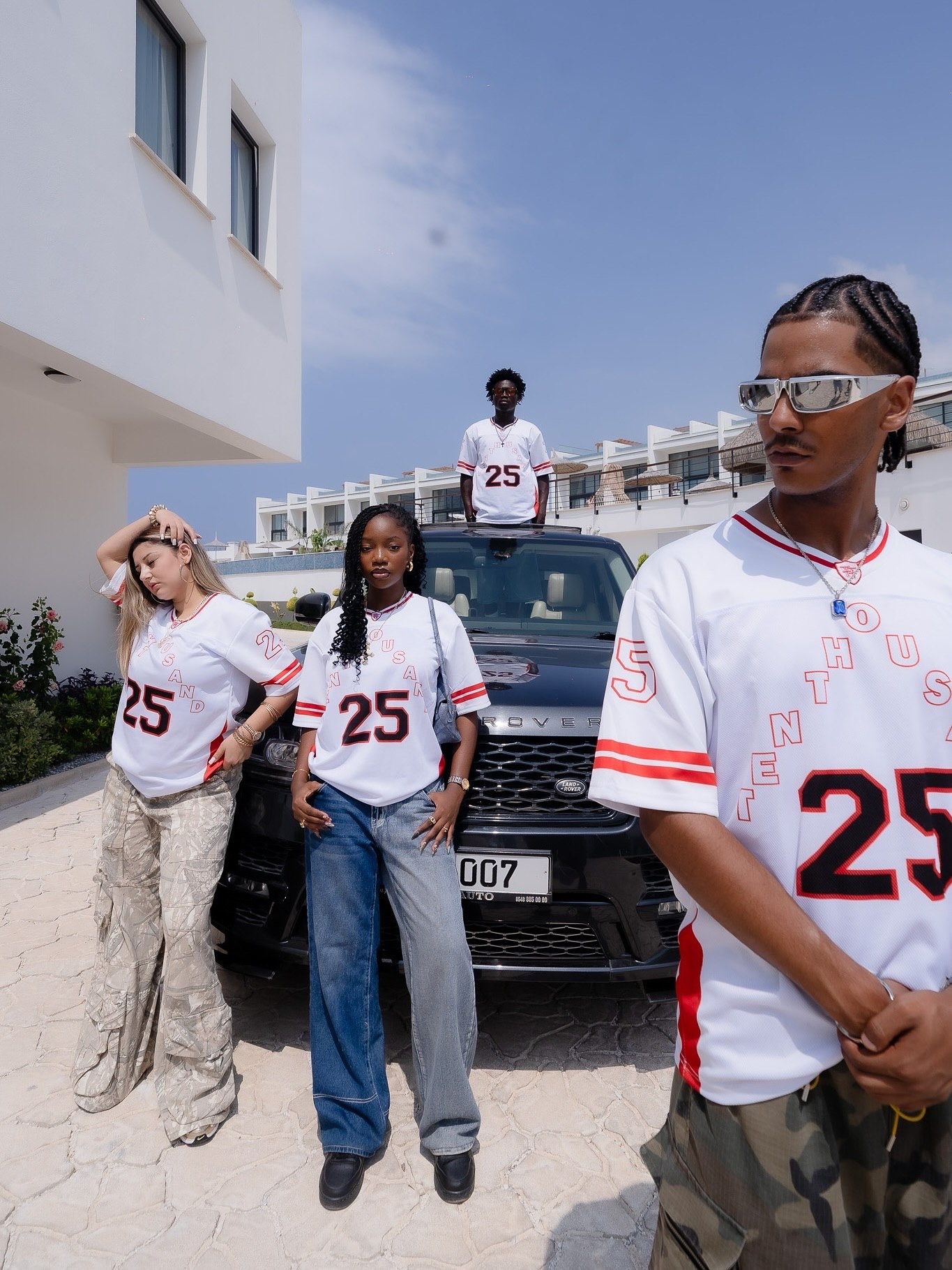 Four people wearing matching white number 25 jerseys posing near a black Range Rover outside a modern white building.