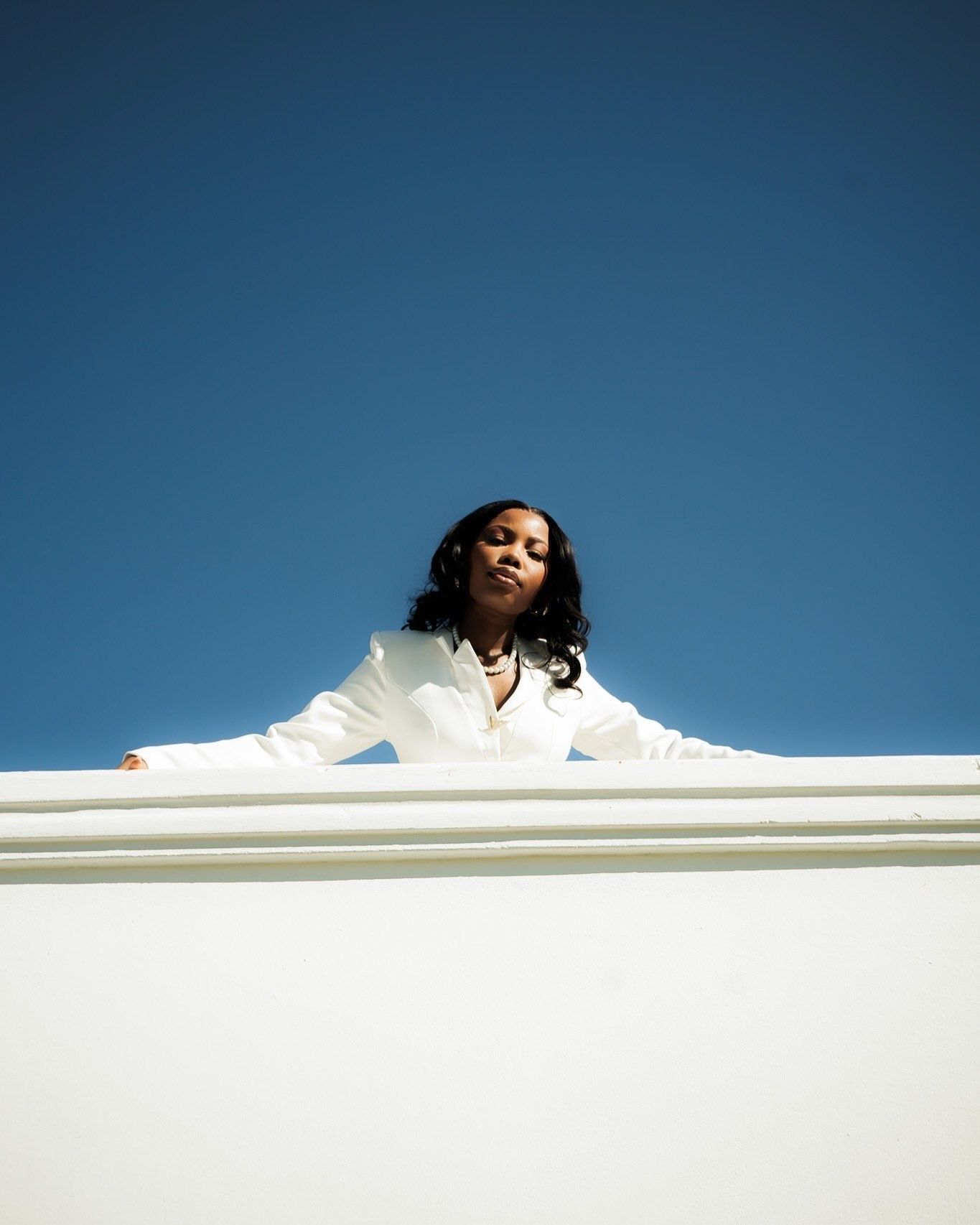 A woman in a white outfit leans against a white wall under a clear blue sky in bright sunlight.