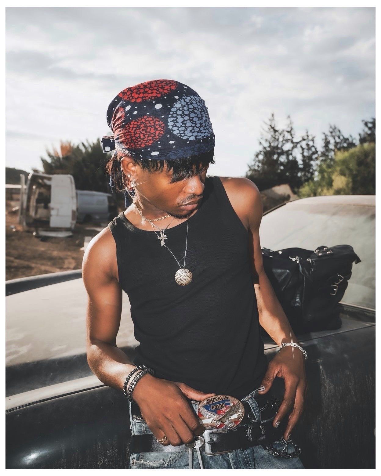 Young person in black tank top, colorful head wrap, and chain necklace leaning on a truck outdoors.