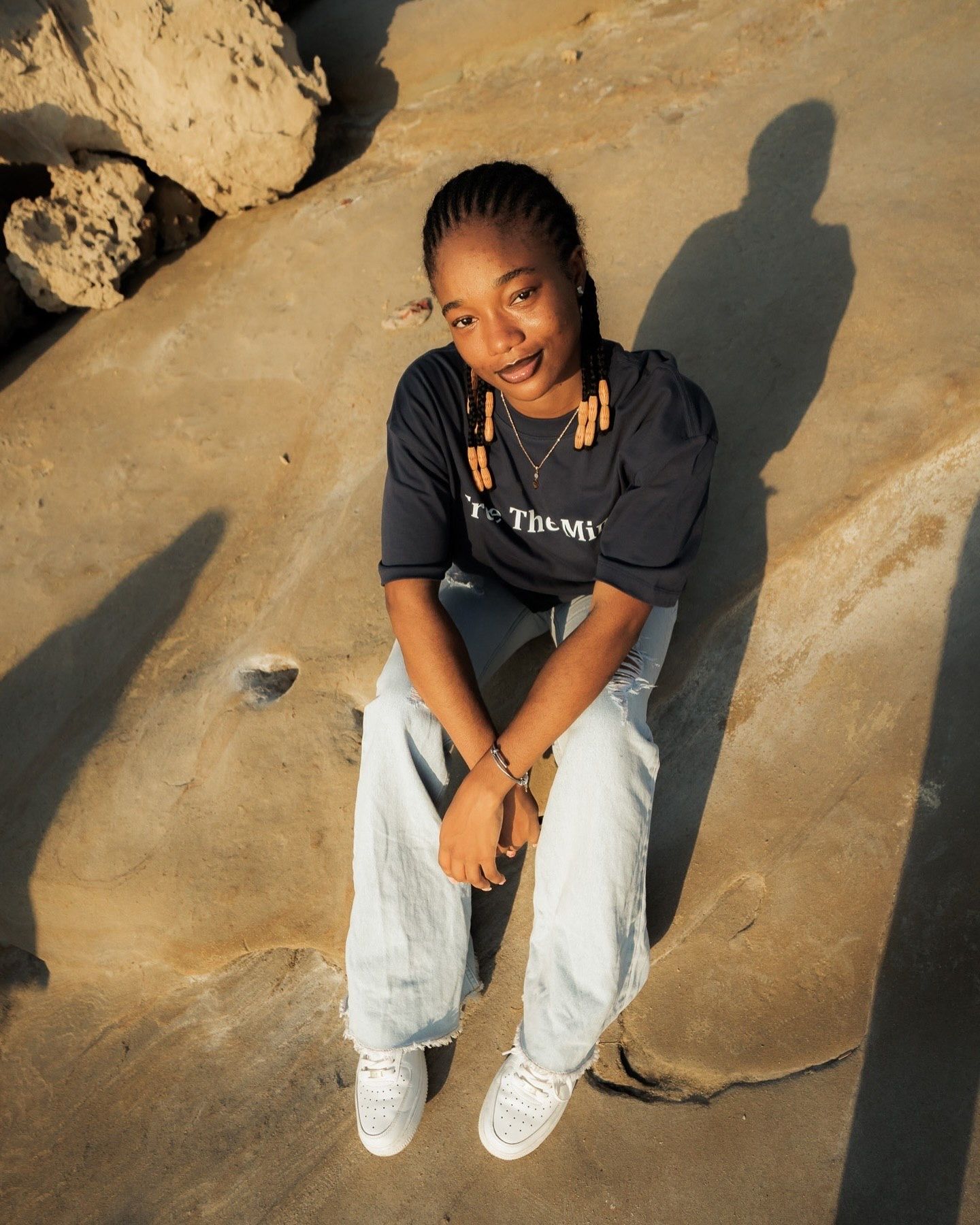 Young woman in black hoodie and light jeans sitting on rocks outdoors, smiling at camera in golden light.