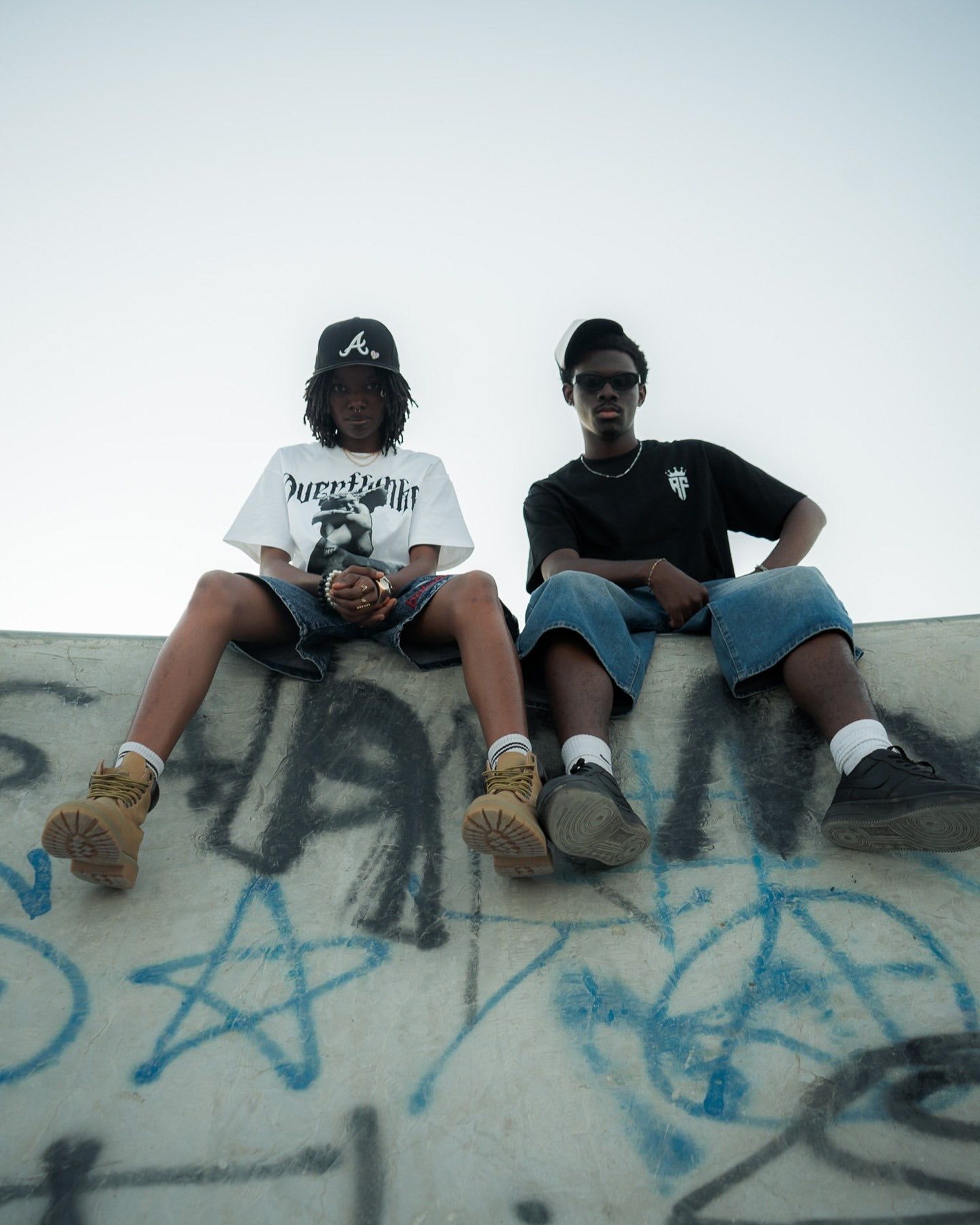 Two young men sitting on a graffiti-covered concrete wall wearing streetwear and caps.