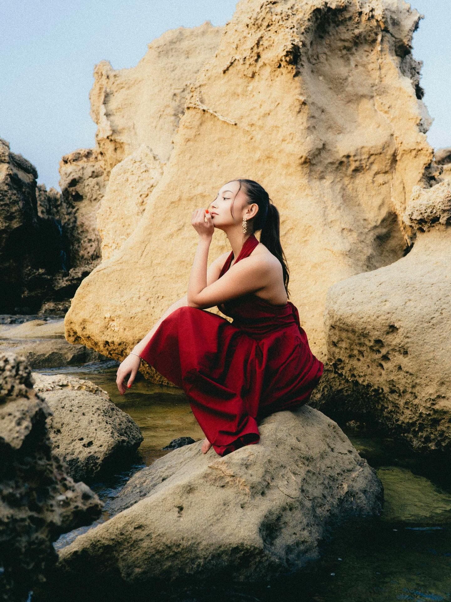 A woman in a red dress sits gracefully on coastal rocks near water, with large sandstone cliffs behind her.