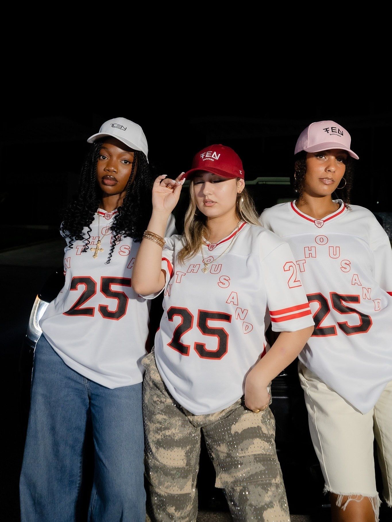 Three women wearing matching white number 25 baseball jerseys and YEN caps pose together at night.