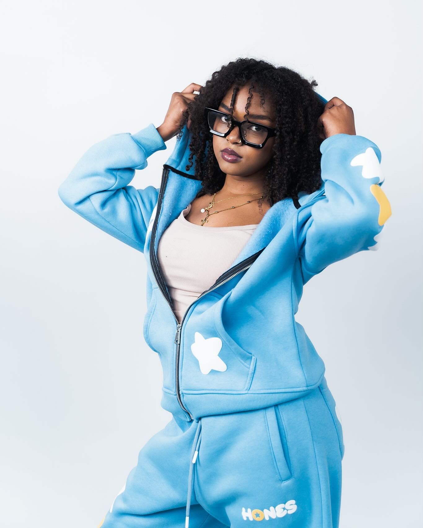 Young woman posing in a light blue HONCS tracksuit with bear patches, wearing black glasses against a white background.