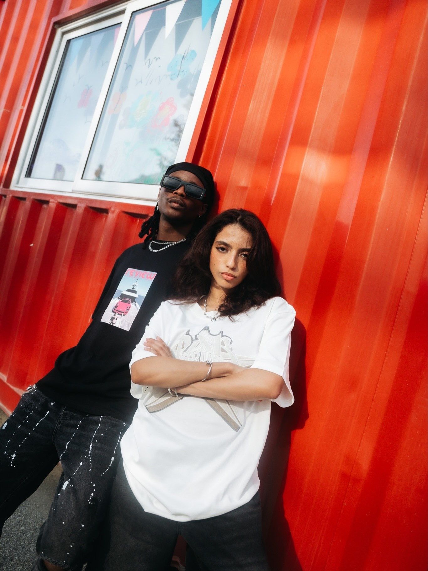 Two young people posing against a vibrant red wall, one in black and one in white oversized tee.