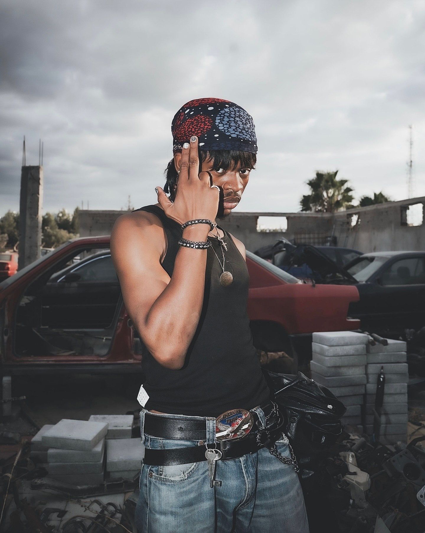 Young man in bandana and tank top poses at a junkyard with cars in background.