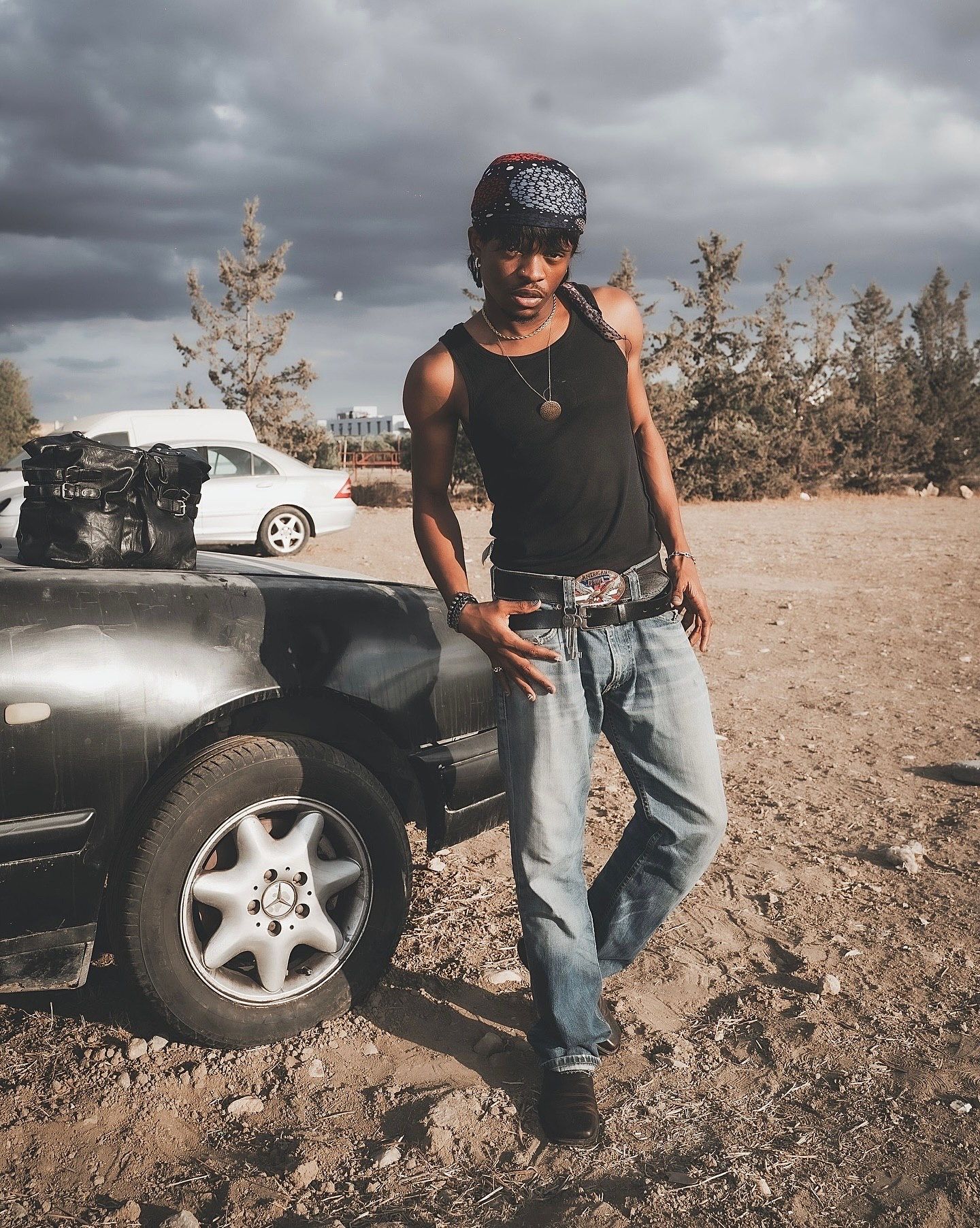 Young man in black tank top, jeans, and bandana leans against a dark SUV on a dirt road under cloudy skies.