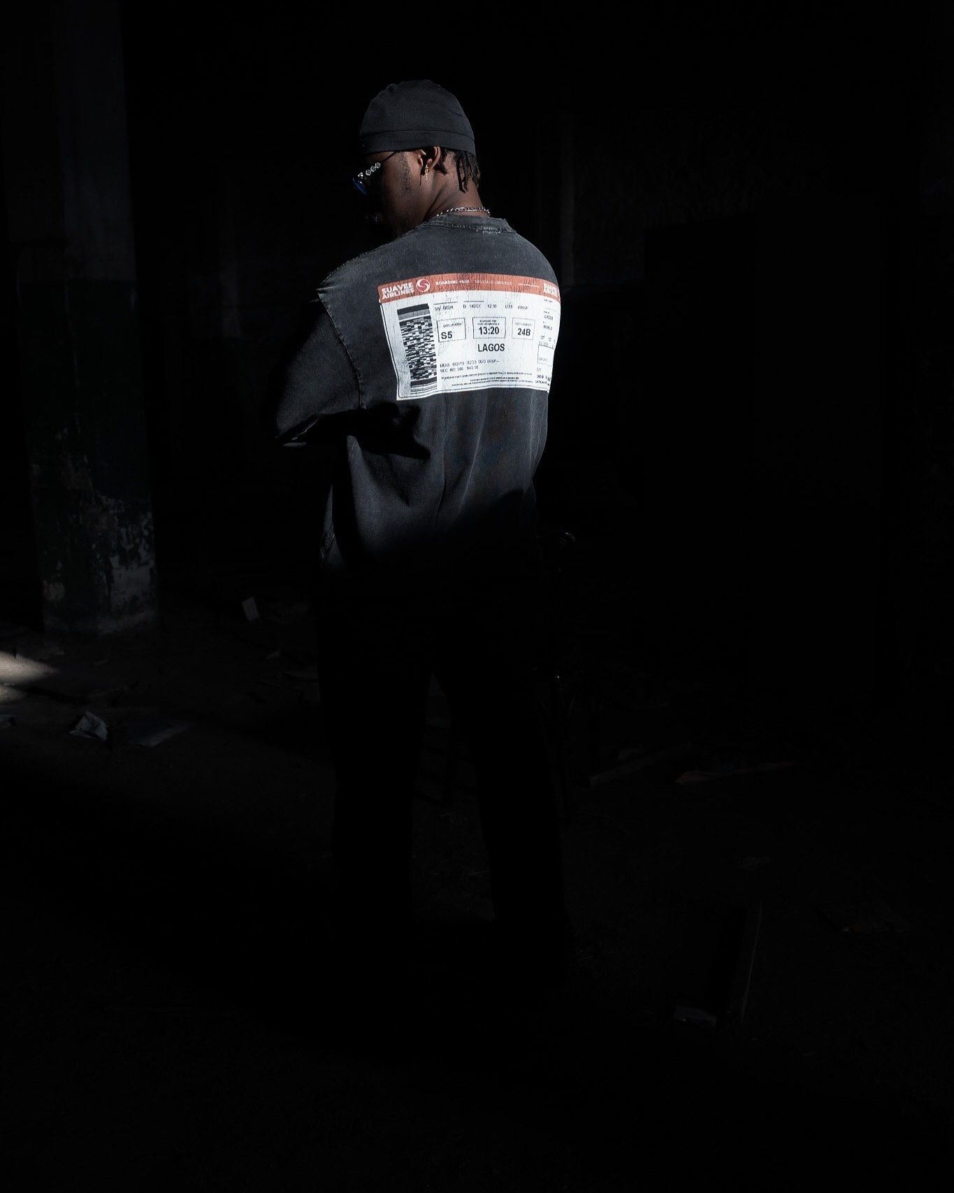Man standing in dark setting wearing gray oversized sweatshirt with label graphic print on back and black cap.