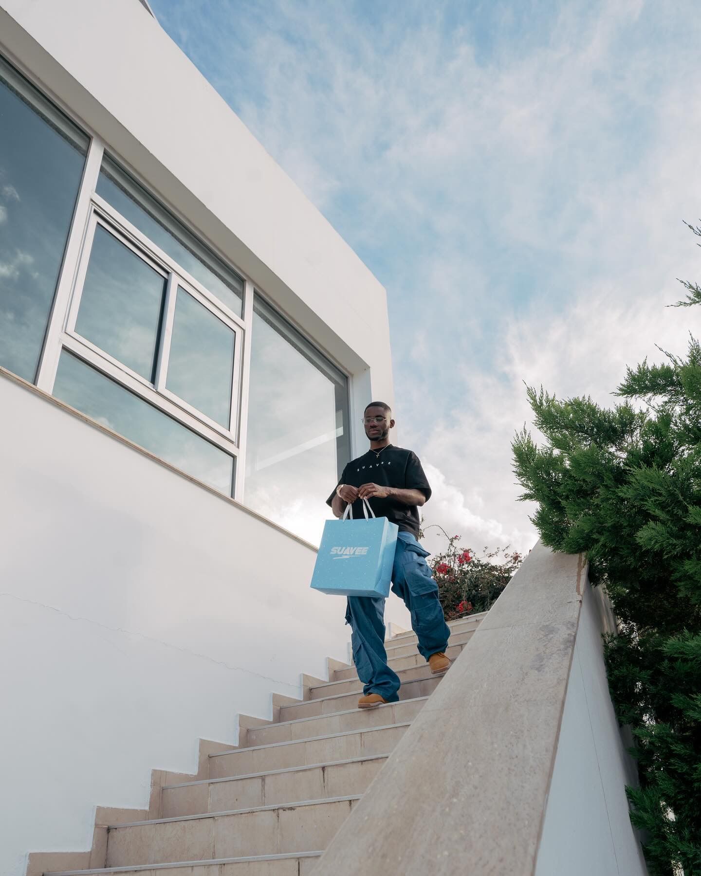 Man descending outdoor stairs holding a light blue shopping bag beside a modern white building with green trees.