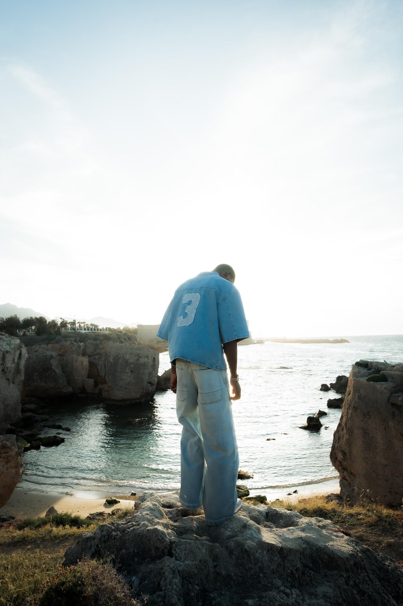 Man in blue shirt and jeans standing on coastal rocks overlooking a scenic ocean cove at sunset.