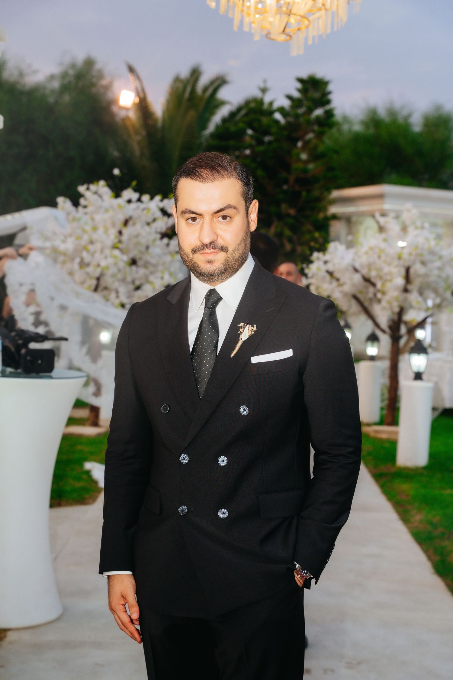 Elegant groom in black double-breasted suit with white boutonniere poses at outdoor wedding venue.