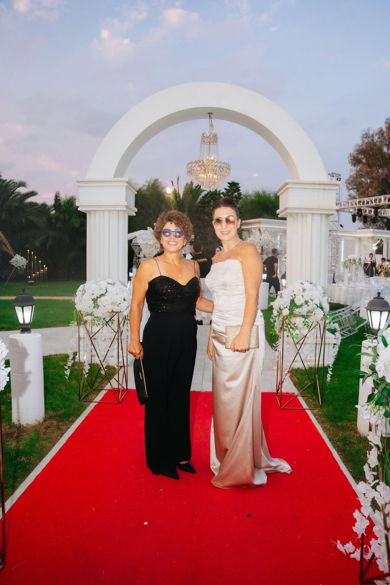 Two elegantly dressed women pose on a red carpet under a white arch at a formal outdoor event.